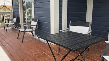 A covered outdoor patio with a table and chairs, wooden flooring, and a dark-colored building with white trim.