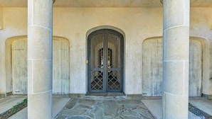 Une entrée d’immeuble avec trois colonnes, une porte centrale ornée d’une grille décorative et un espace d’entrée pavé de pierres.
