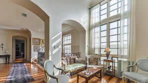 Un salon spacieux doté d’un parquet en bois, d’une grande fenêtre avec des rideaux et d’un coin salon comprenant un canapé et des fauteuils.