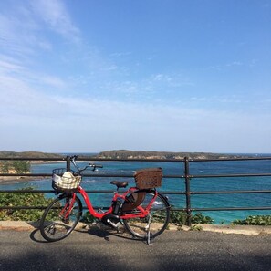 A red bicycle with a basket and a brown basket attached to the back, parked on a paved area with a view of the sea and distant hills. - Midori Guest House (Tsushima)
