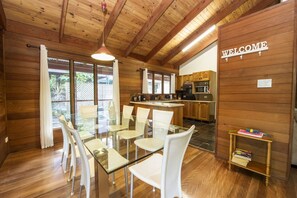 Un intérieur en bois avec une table et des chaises en verre pour la salle à manger, un coin cuisine et une pancarte « BIENVENUE ».
