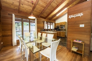 A wooden interior with a glass dining table and chairs, a kitchen area, and a 'WELCOME' sign.