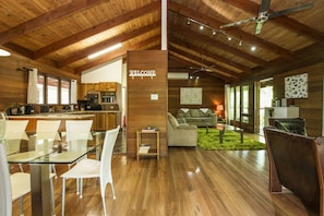A spacious living area with a glass dining table, white chairs, a sofa, and a coffee table. A 'WELCOME' sign is visible on the wall.