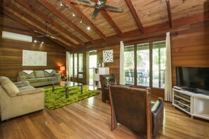 A wooden-paneled room with a ceiling fan, a sofa, a coffee table, a TV, and large windows.