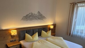 A hotel room with a bed, wooden headboard, two bedside lamps, a nightstand, a wall-mounted lamp, and a window with curtains. - Apartment for 1-2 people, separate bedroom, shower / toilet, balcony, 40 sqm (Schönau am Königssee)