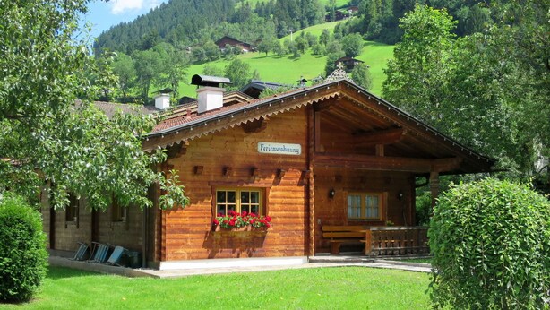 A wooden chalet with a sign reading "Ferienwohnung" in front, surrounded by lush greenery and hills in the background.