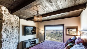 Ein Schlafzimmer mit Stehwänden, einem Bett mit bunten Kissen, einem Fernseher und einem großen Fenster mit Blick ins Grüne.