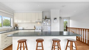 A modern kitchen with a large island, white cabinetry, stainless steel appliances, and a wooden bar stool seating area. - The Beach Hive - Inlet Side Inverloch (INVERLOCH)