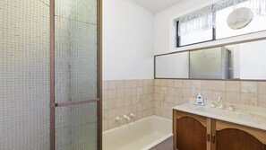 A bathroom with a bathtub, a wooden cabinet, a mirror, and a window with lace curtains. - The Beach Hive - Inlet Side Inverloch (INVERLOCH)