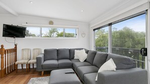 A modern living room with a grey sectional sofa, a flat-screen TV, and a balcony with a view of trees. - The Beach Hive - Inlet Side Inverloch (INVERLOCH)