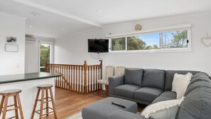 A modern living room with a grey sofa, wooden bar stools, a flat-screen TV mounted on the wall, and a large window offering a view of trees. - The Beach Hive - Inlet Side Inverloch (INVERLOCH)