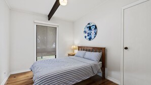 A bedroom with a bed, a window with a view, a nightstand, and a wall art piece. - The Beach Hive - Inlet Side Inverloch (INVERLOCH)