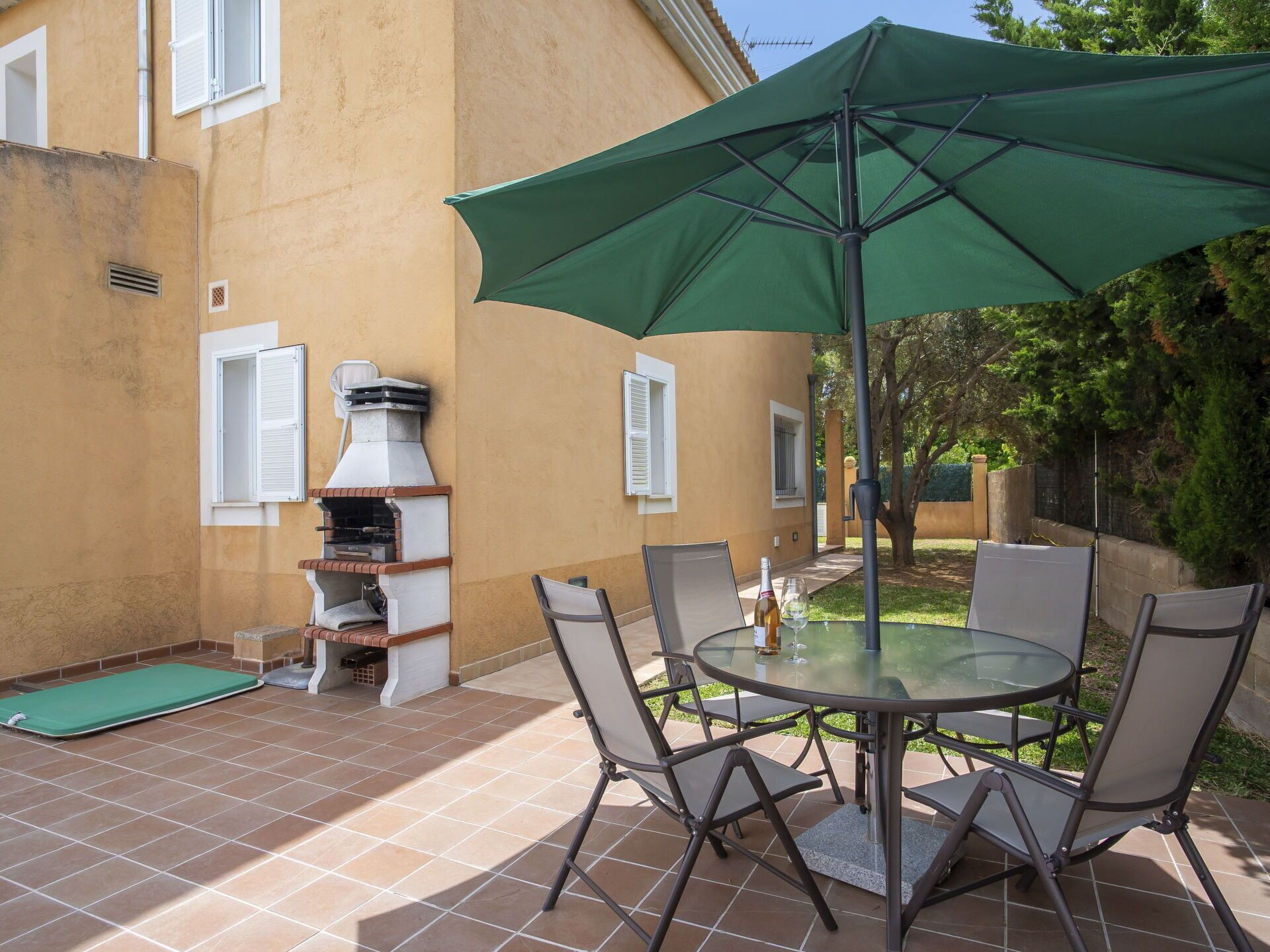 An outdoor patio with a round glass table, six chairs, a green umbrella, and a brick oven.