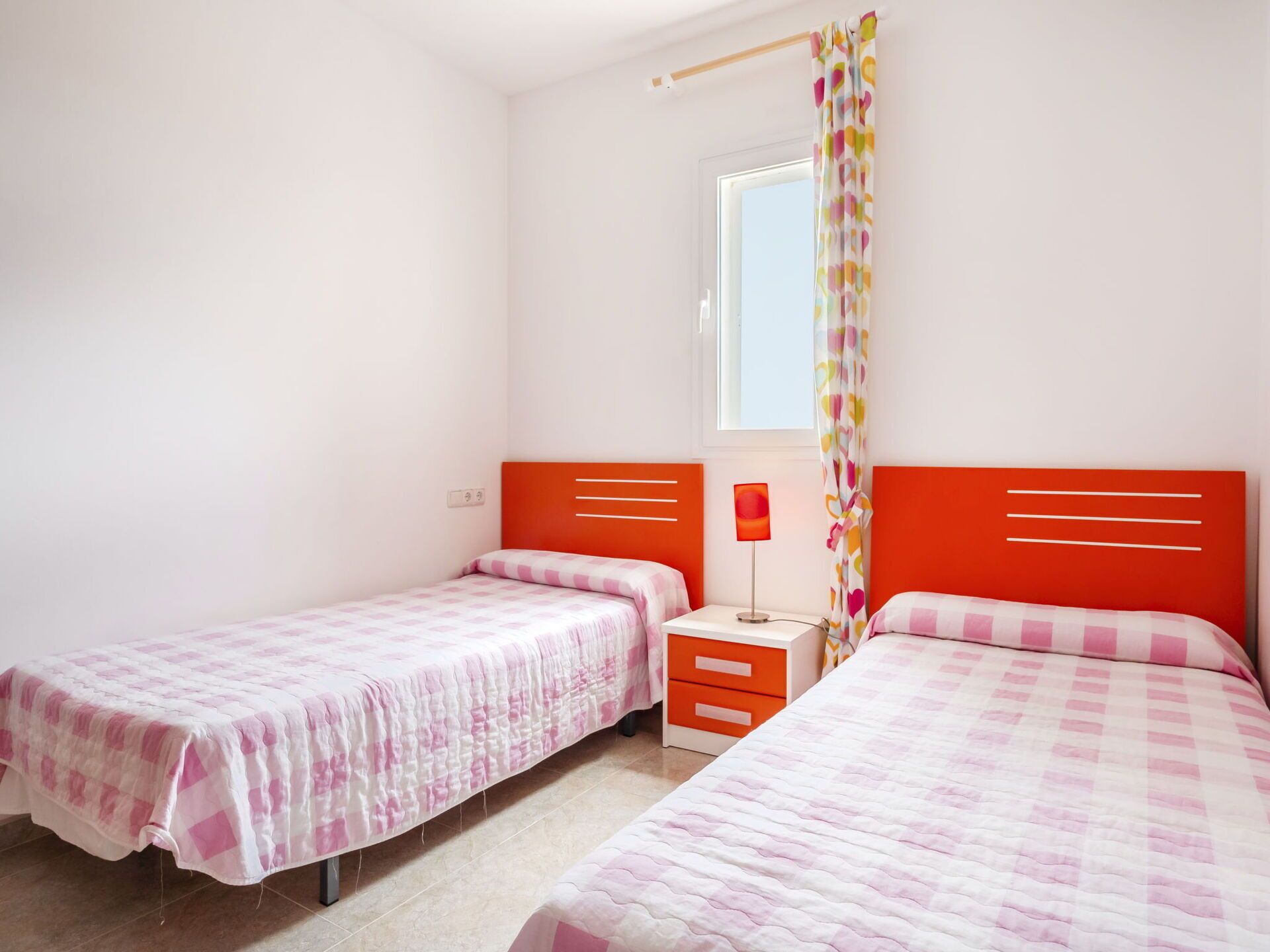 Een slaapkamer met twee enkele bedden, een nachtkastje, een lamp en een raam met gordijn.