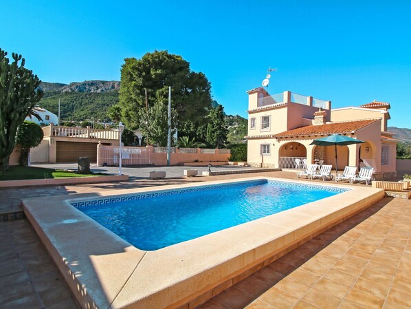 A swimming pool with a tiled deck, a patio area with lounge chairs, and a building with a tiled roof and a satellite dish.