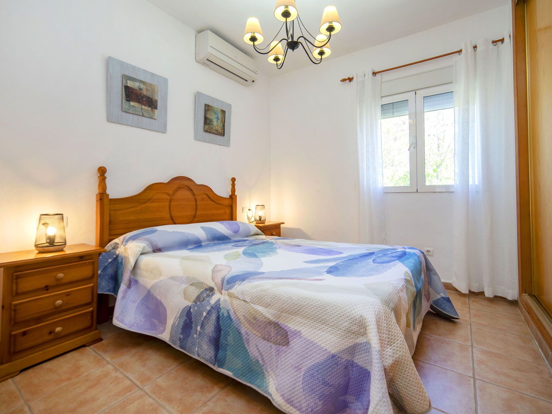 A bedroom with a wooden bed, two bedside tables, a chandelier, an air conditioner, and a window with curtains.