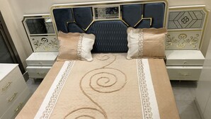 Una cama con colcha color crema y adornos en espiral, con dos mesitas de noche con cajones y un tocador con espejo.