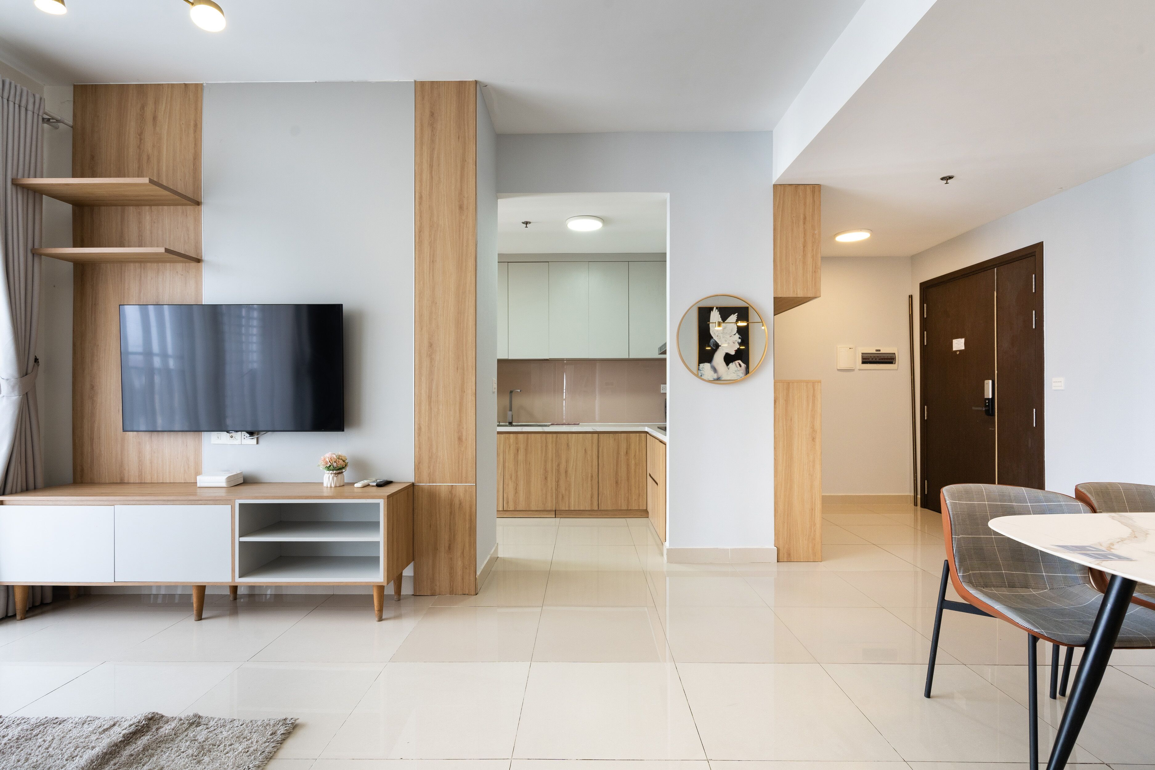 A modern living room with a flat-screen TV, wooden shelving, a dining area with a table and chairs, and a kitchen in the background.