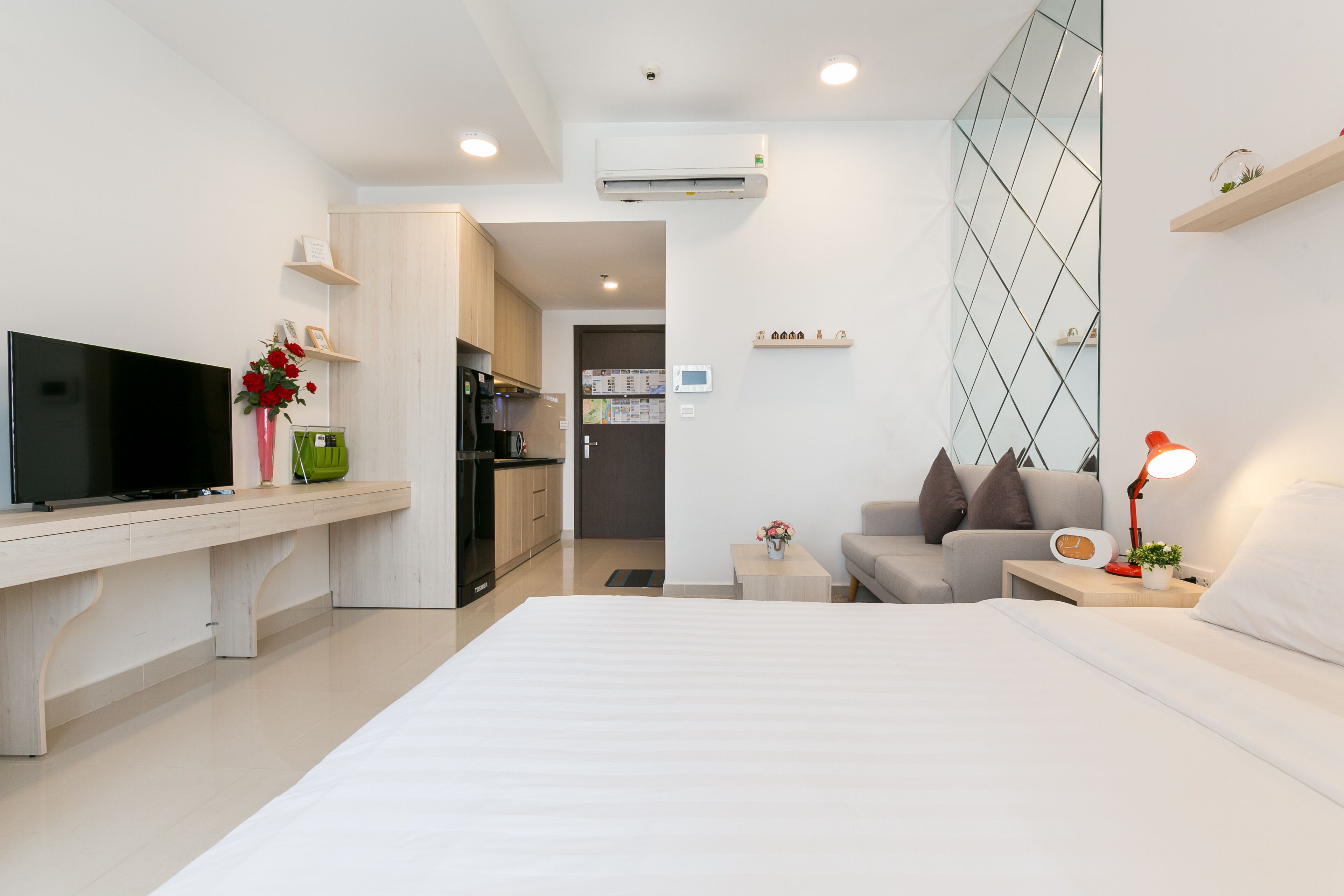 A modern hotel room with a flat-screen TV, a dining area, a kitchenette, and a cozy seating area with a lamp.