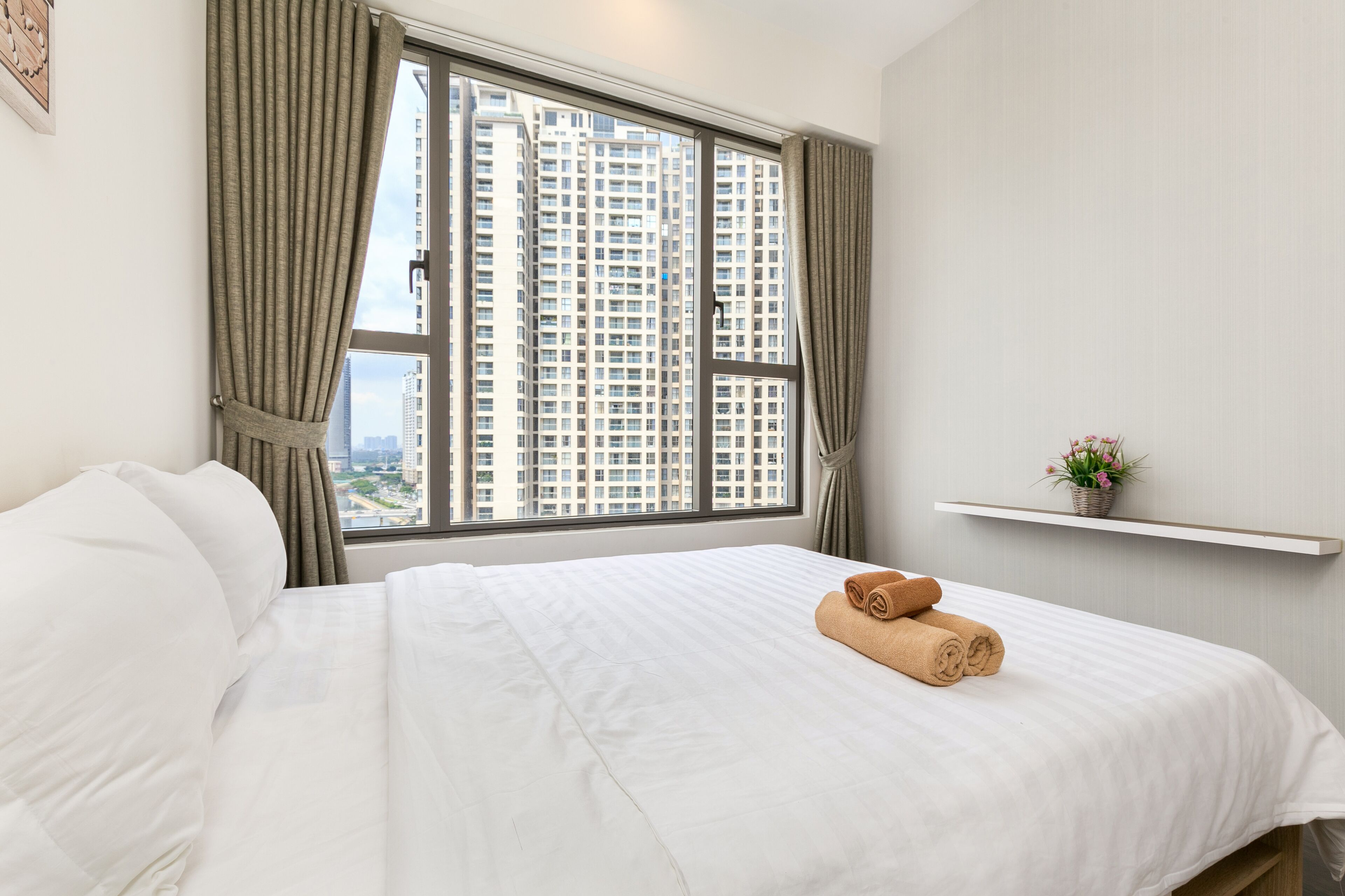 A hotel room with a large window, a neatly made bed, and a small shelf with a plant.