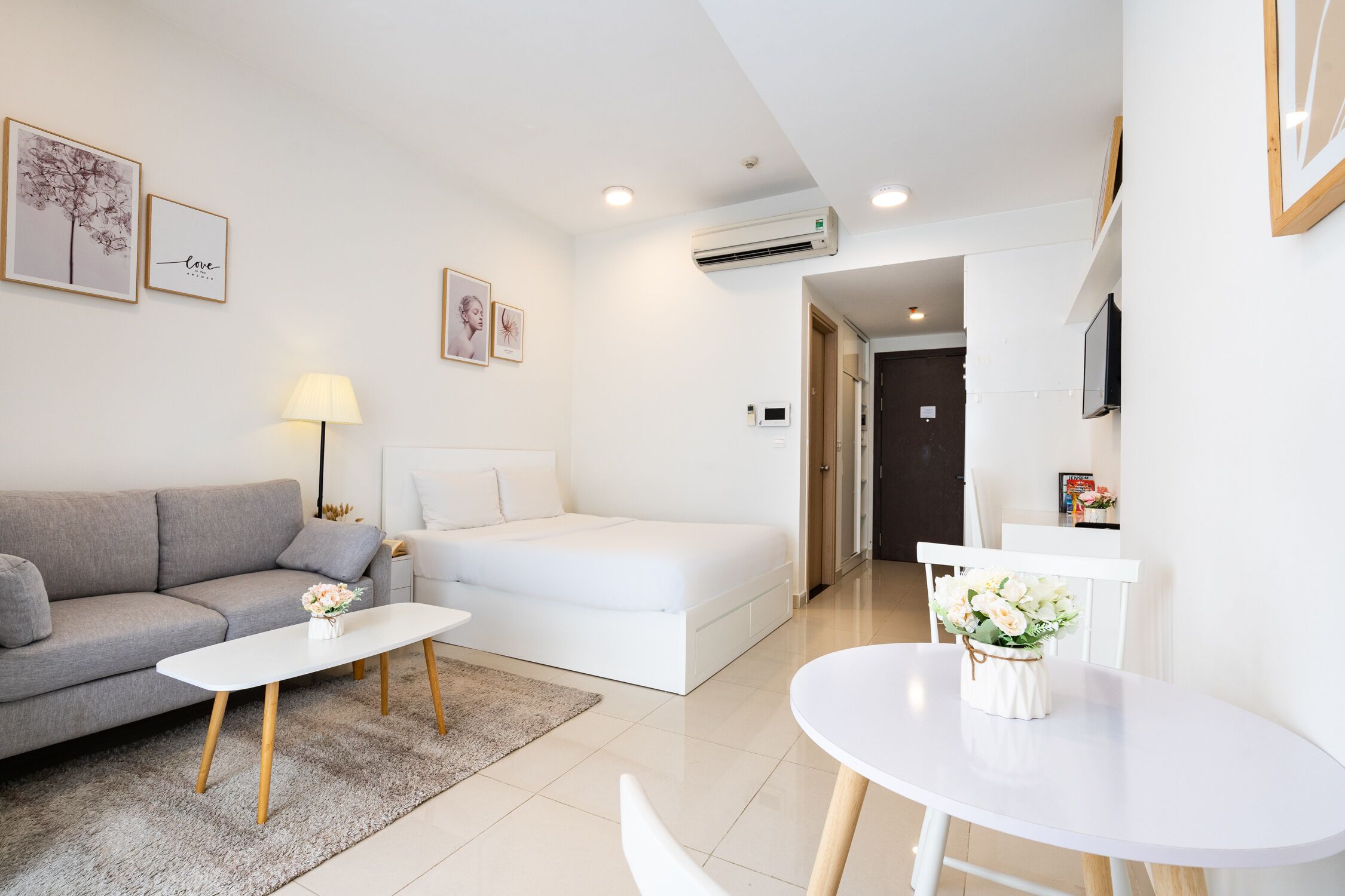 A modern living room with a grey sofa, a white bed, a round table with a vase of flowers, and a flat-screen TV mounted on the wall.