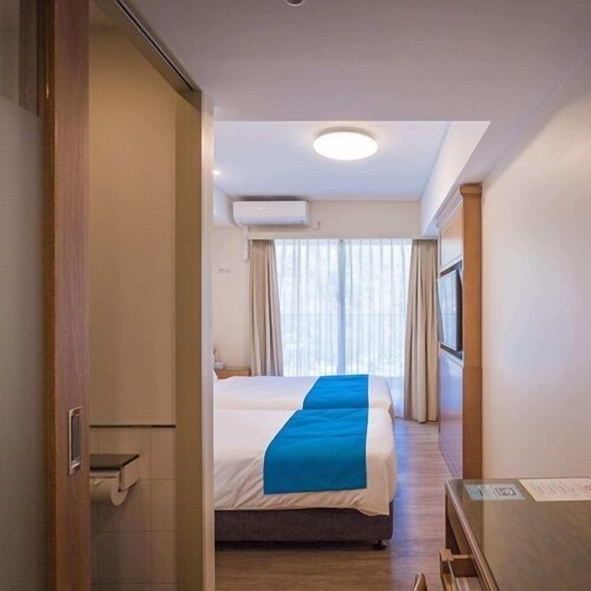 A hotel room with a bed, a desk, a chair, a window with curtains, and an air conditioning unit on the wall.