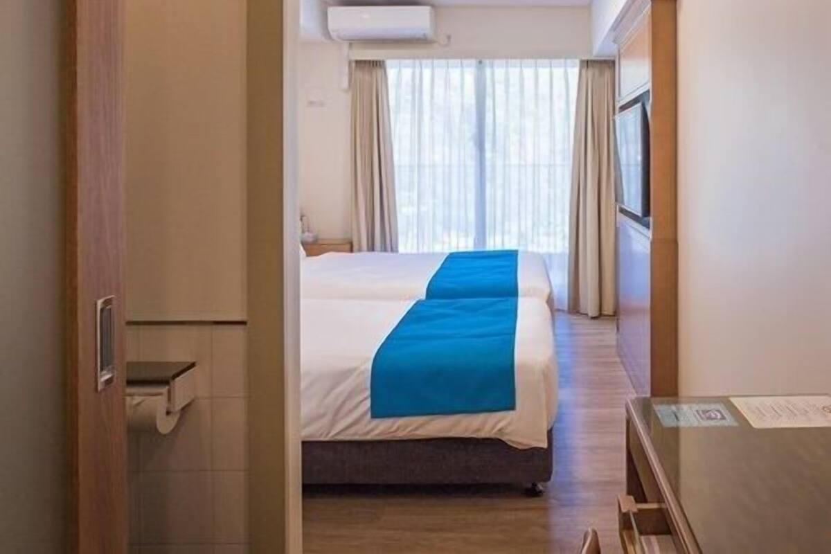 A hotel room with a bed, a desk, a chair, a window with curtains, and an air conditioning unit on the wall.