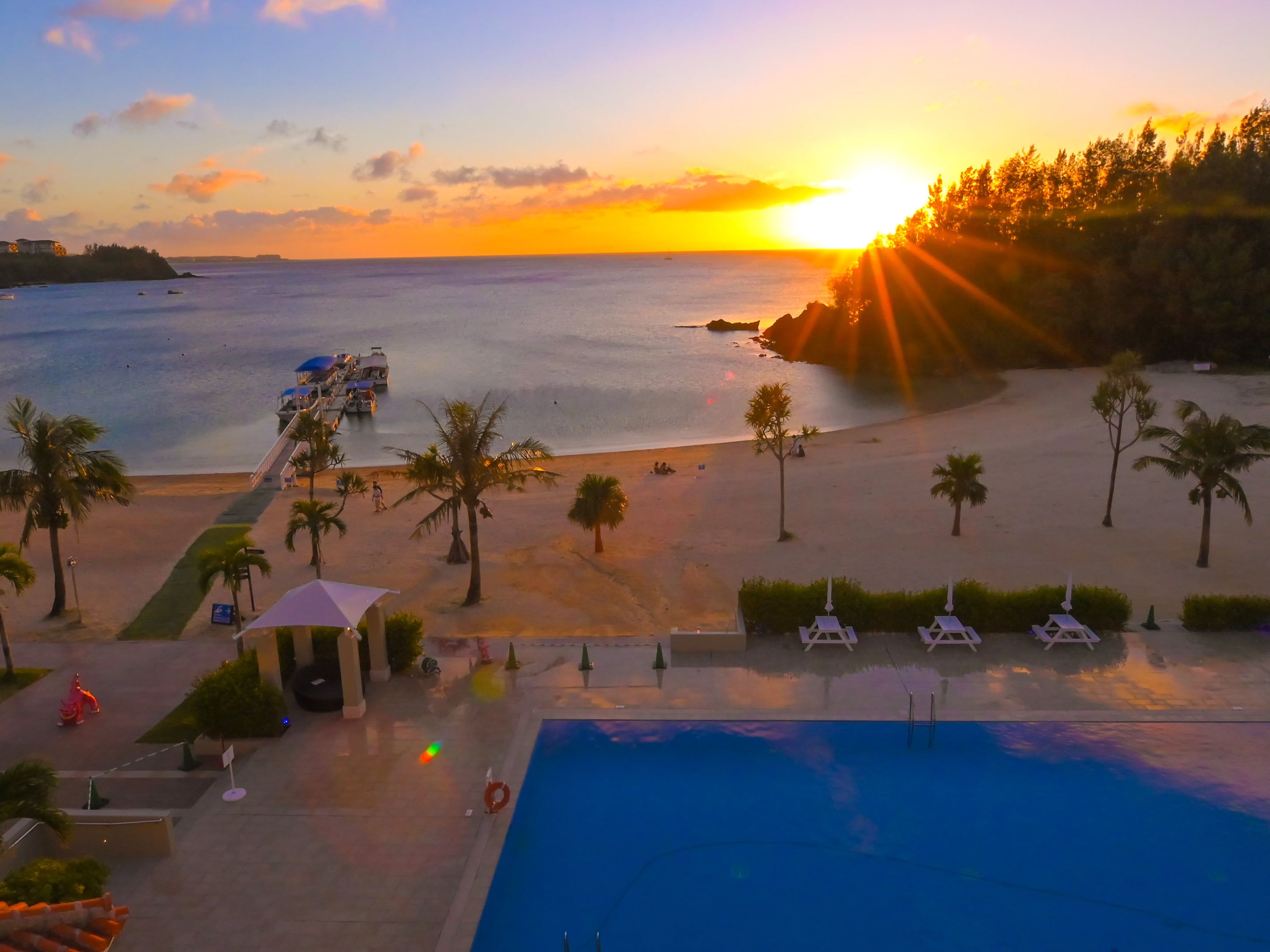 A beach resort with a swimming pool, palm trees, and a sunset view.