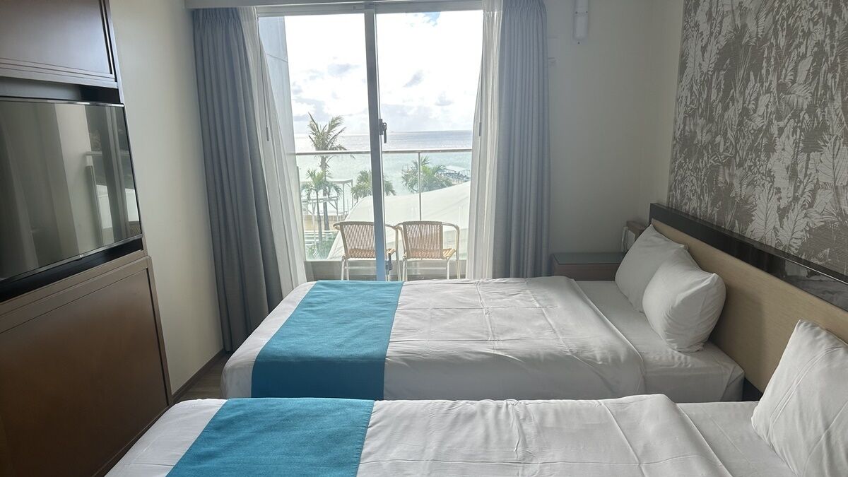 A hotel room with two beds, a TV, and a balcony with a view of the ocean.