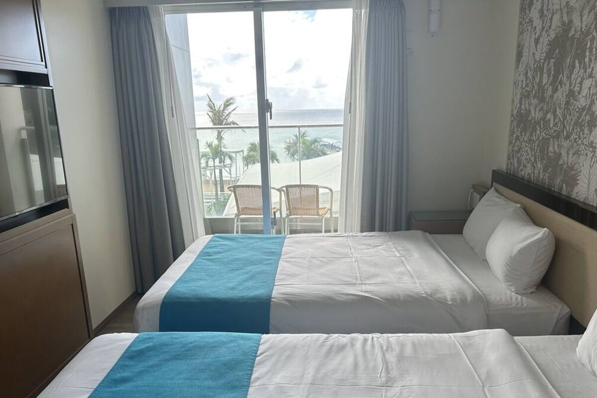 A hotel room with two beds, a TV, and a balcony with a view of the ocean.