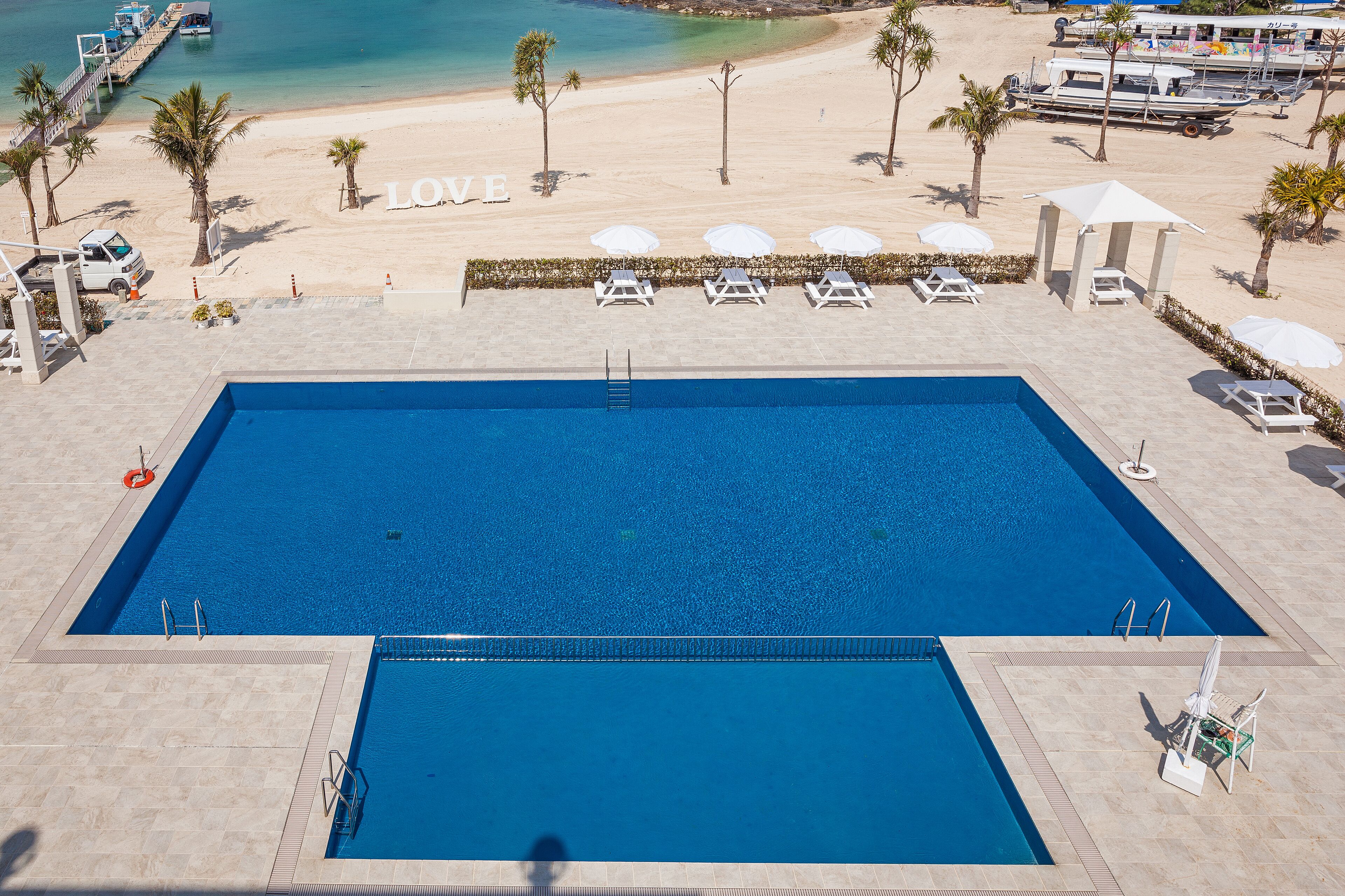 Una piscina con un cartel que dice 'LOVE' en la playa, sillas de descanso y un puesto de salvavidas.