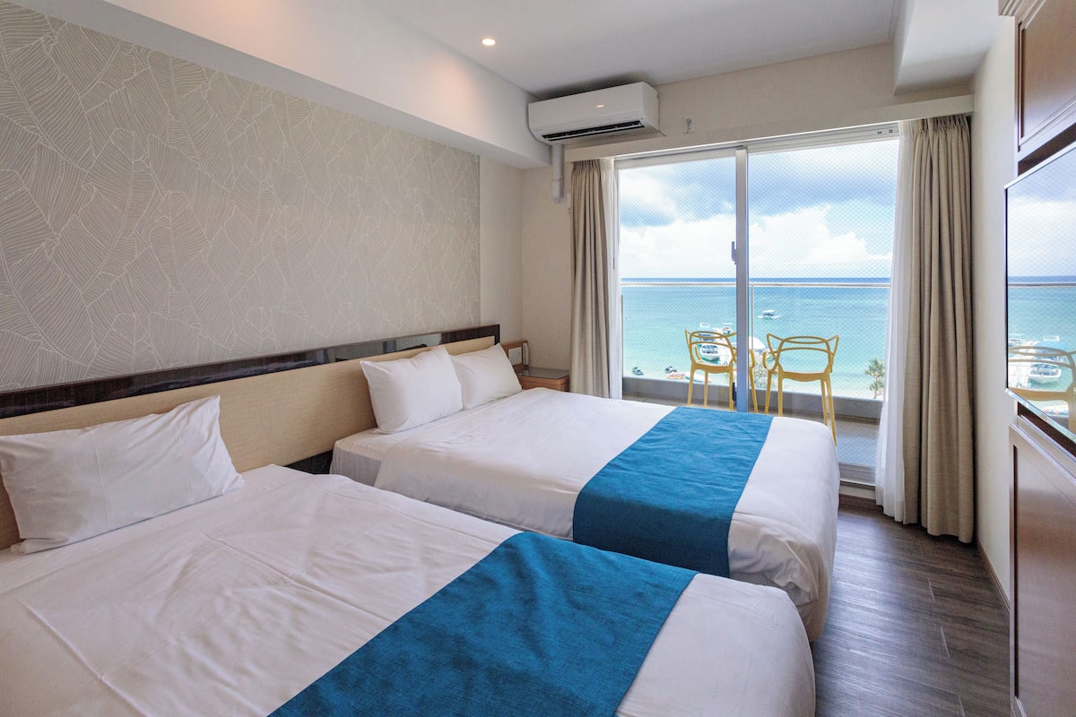 A hotel room with two beds, a balcony with a view of the sea, and a small table and chair.
