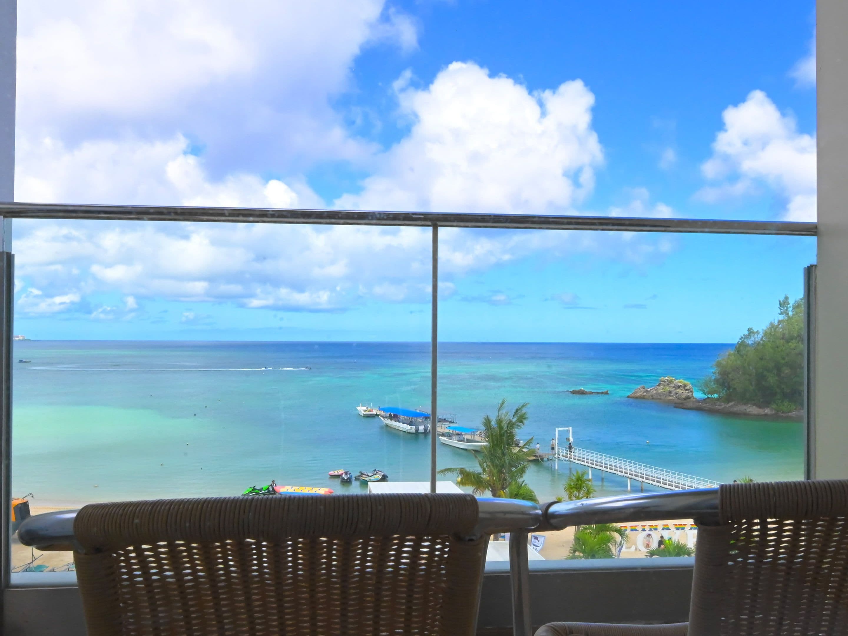 A balcony with wicker chairs overlooking a coastal view, including a pier and boats.