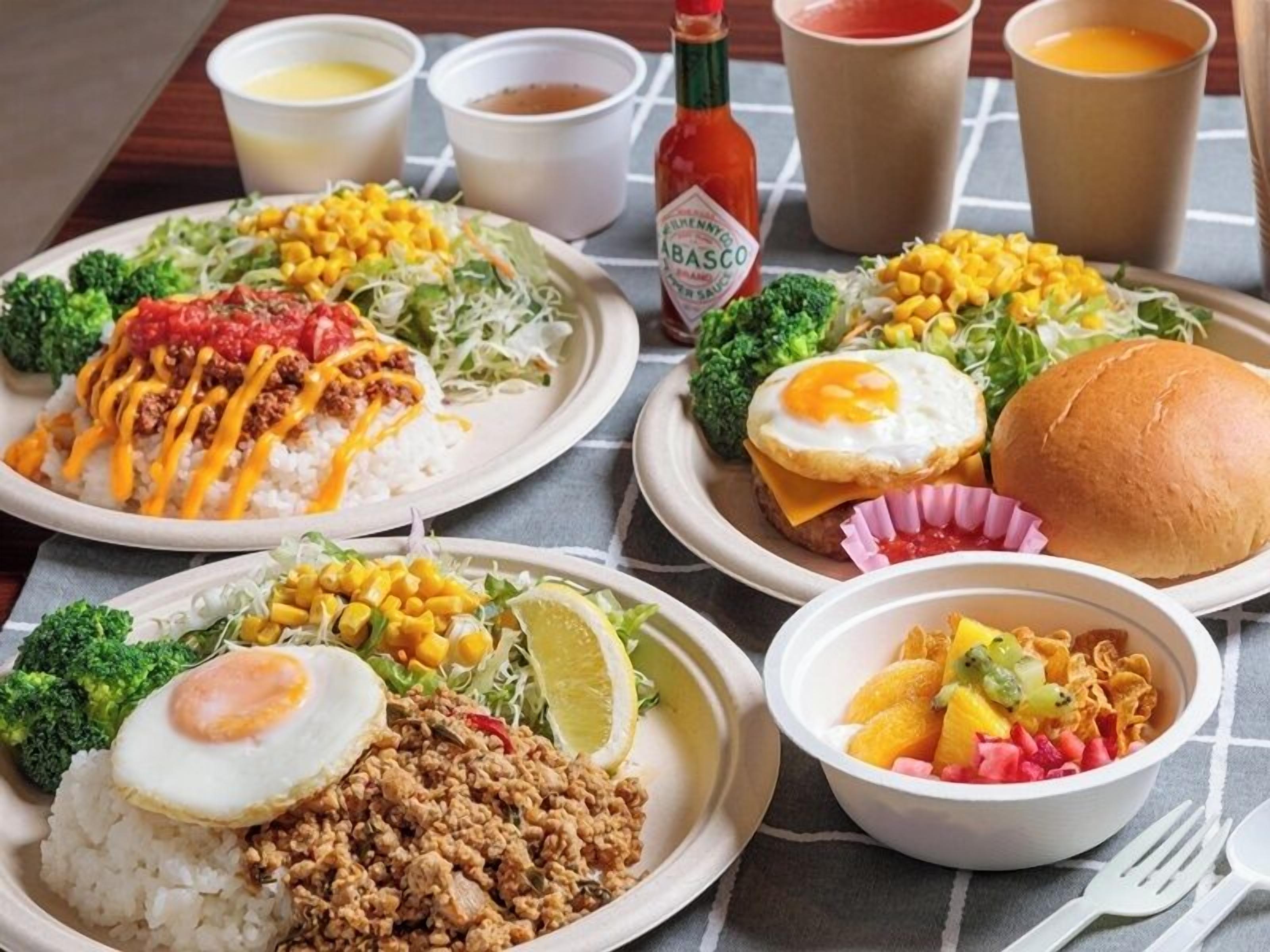 A table with various dishes including rice topped with cheese and meat, a burger, and a bowl of fruit salad.