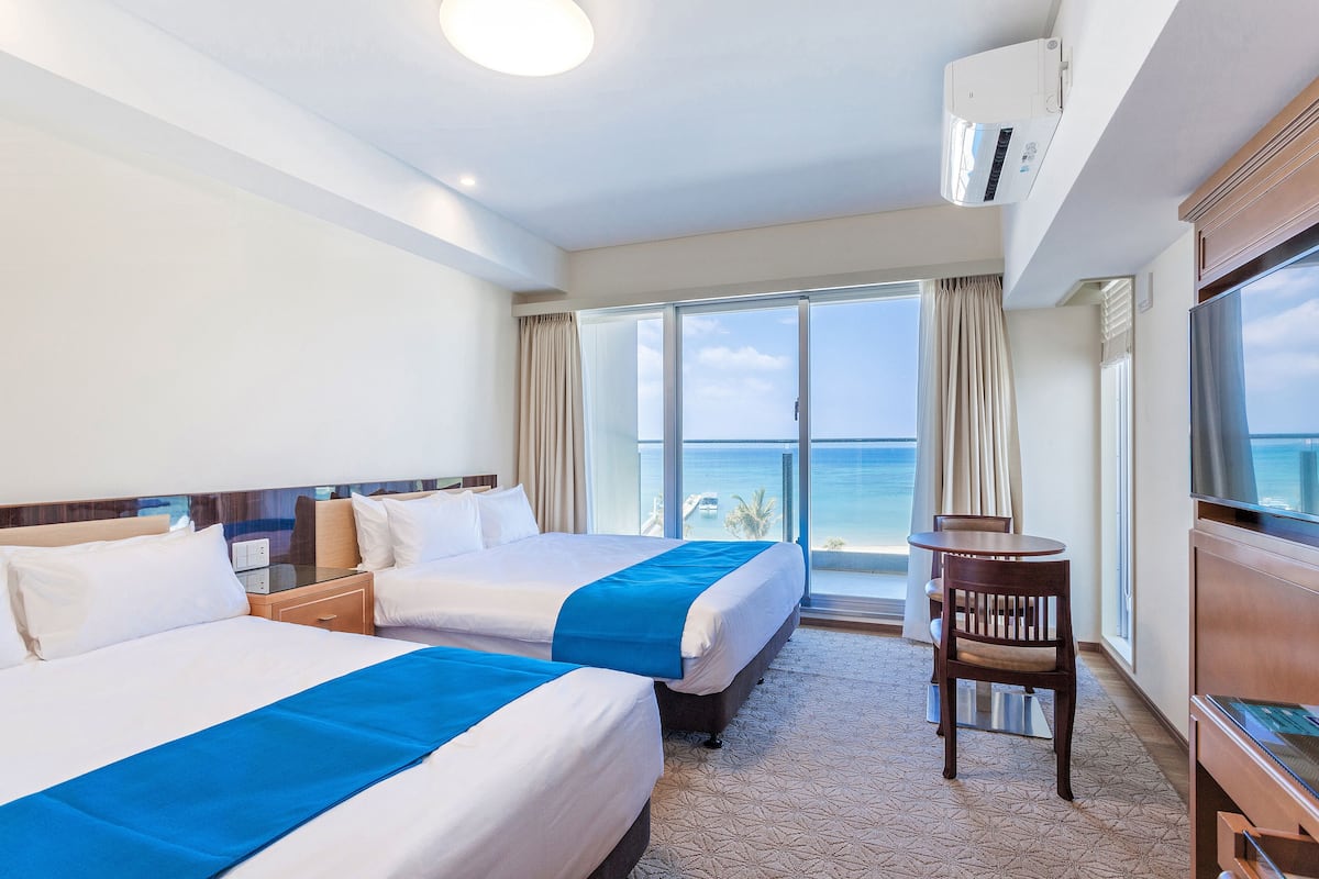 A hotel room with two beds, a TV, a small table, and a view of the ocean.
