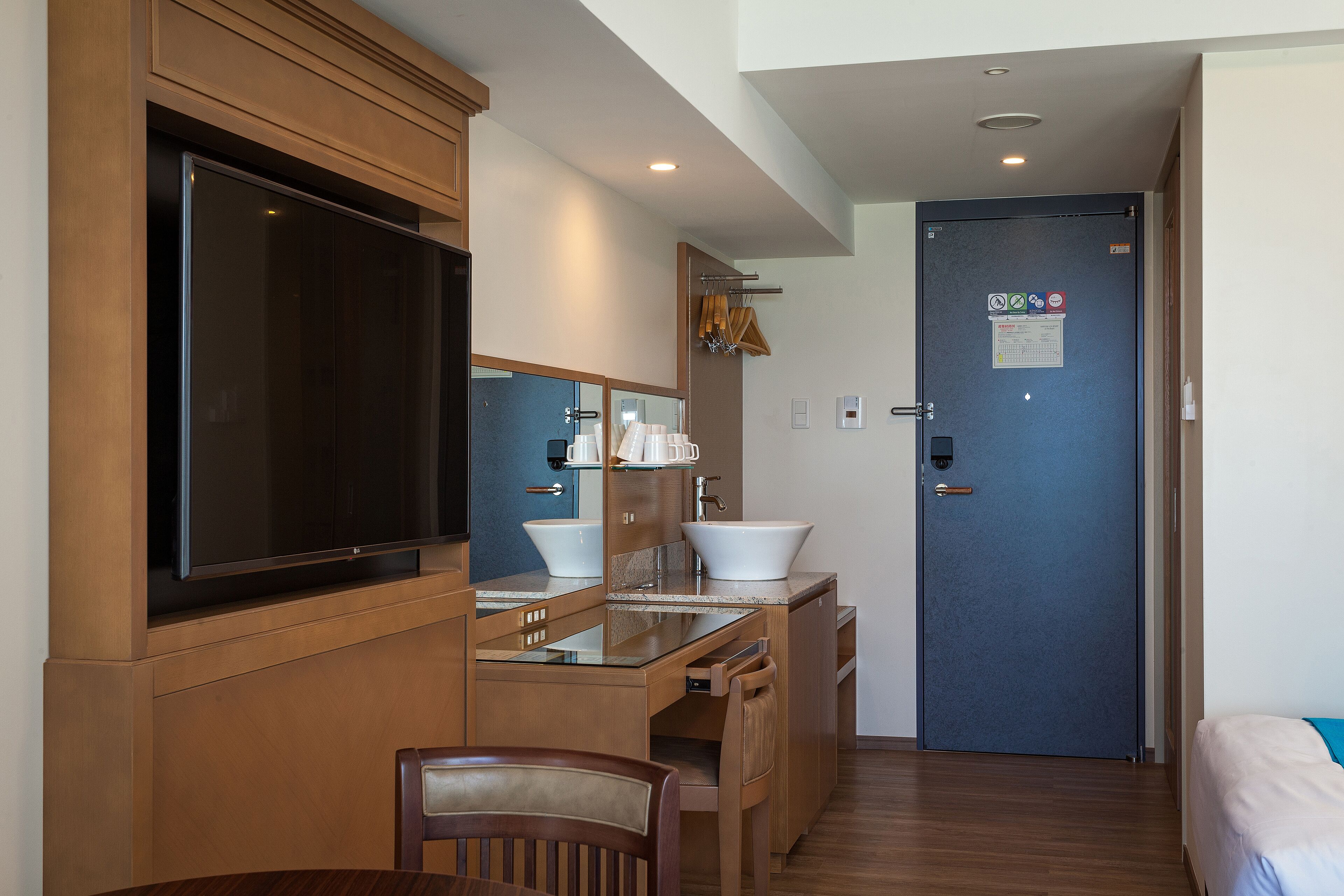 A compact hotel room with a wooden vanity, a large mirror, a sink, and a flat-screen TV.
