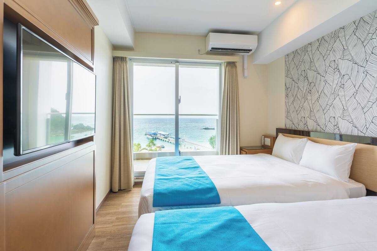 A hotel room with two beds, a large window with a view of the sea, and a patterned wall.