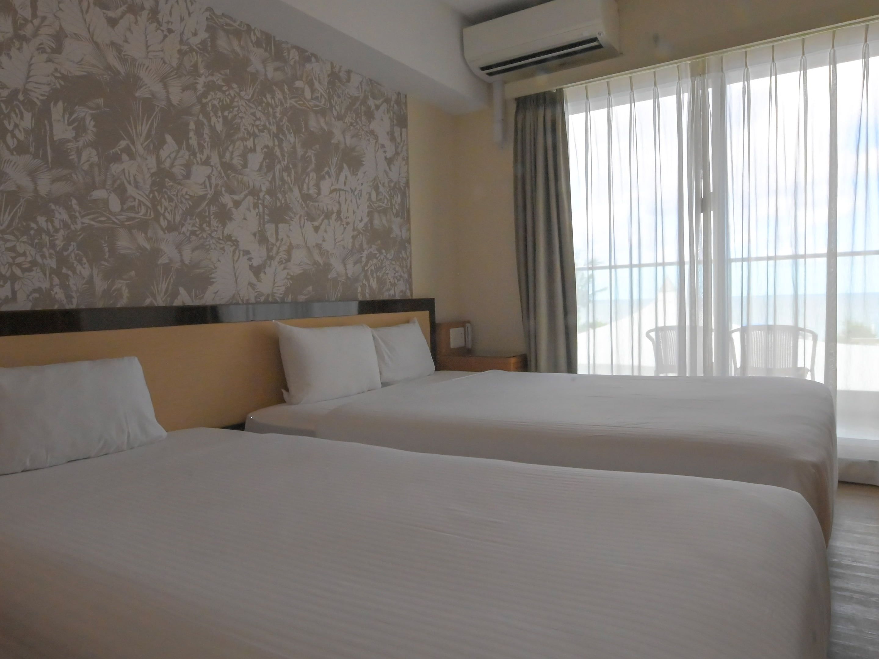 Une chambre d’hôtel avec deux lits, une grande fenêtre avec des rideaux, un appareil de climatisation et un mur orné d’un motif floral.