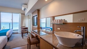 Una habitación de hotel con un ventanal grande, una cama, un escritorio, una silla, un lavamanos y un espejo.