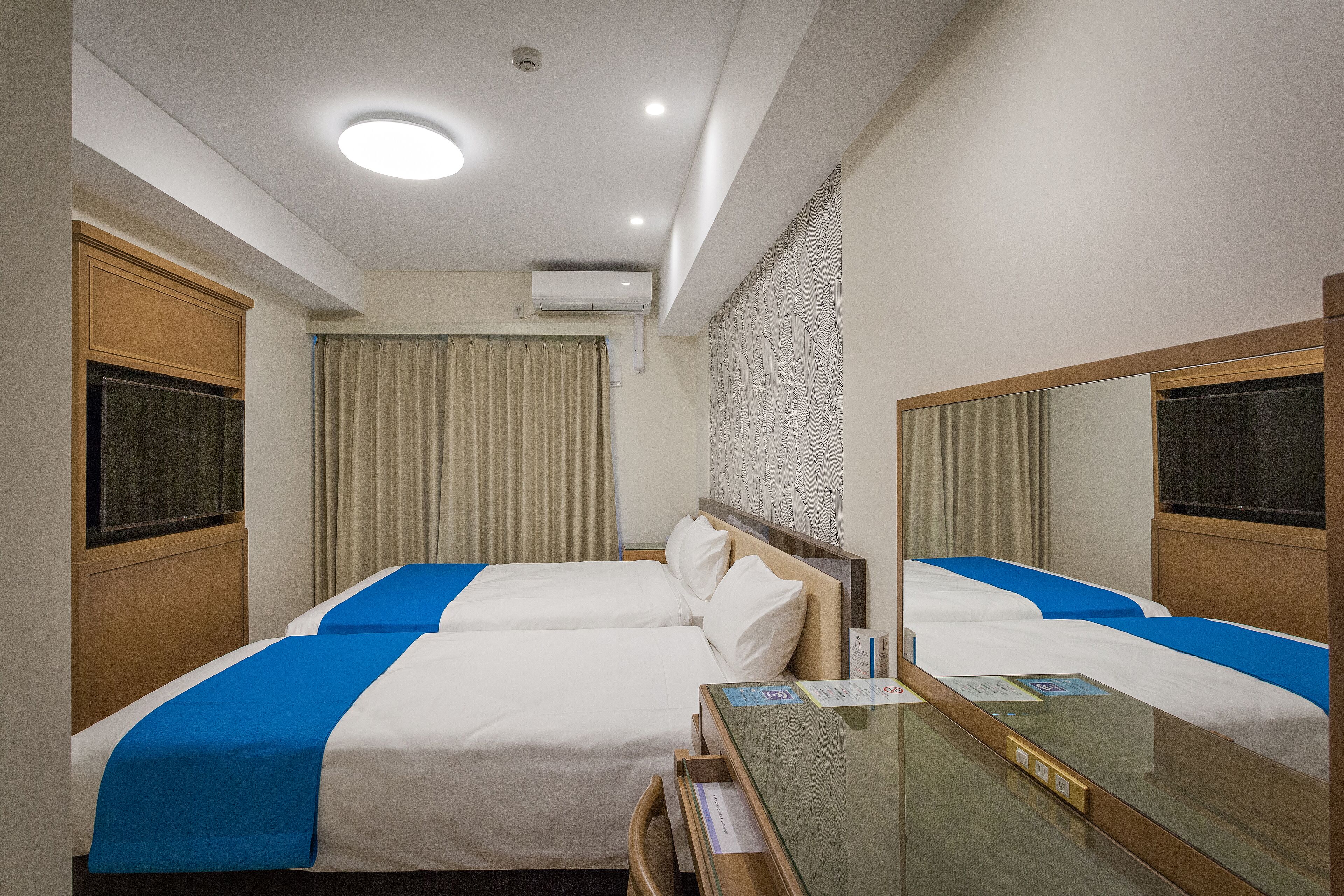 A hotel room with two beds, a desk, a mirror, and a window with curtains.