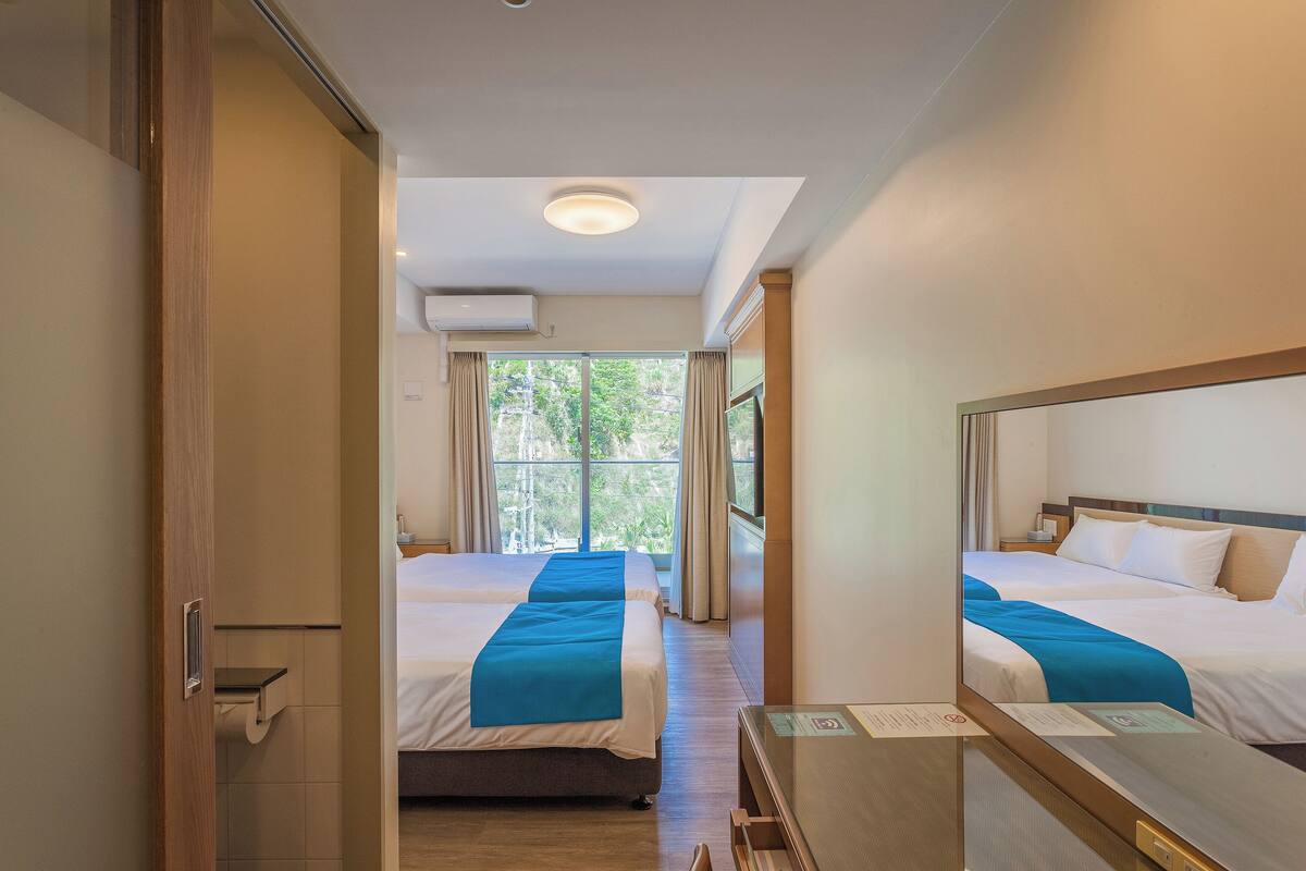 A hotel room with two beds, a wooden floor, a small desk, and a window with curtains.