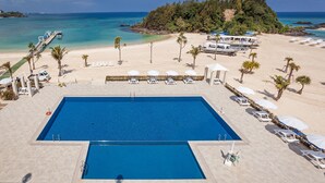 Una piscina con tumbonas y sombrillas, una playa con un muelle, y una isla boscosa al fondo.