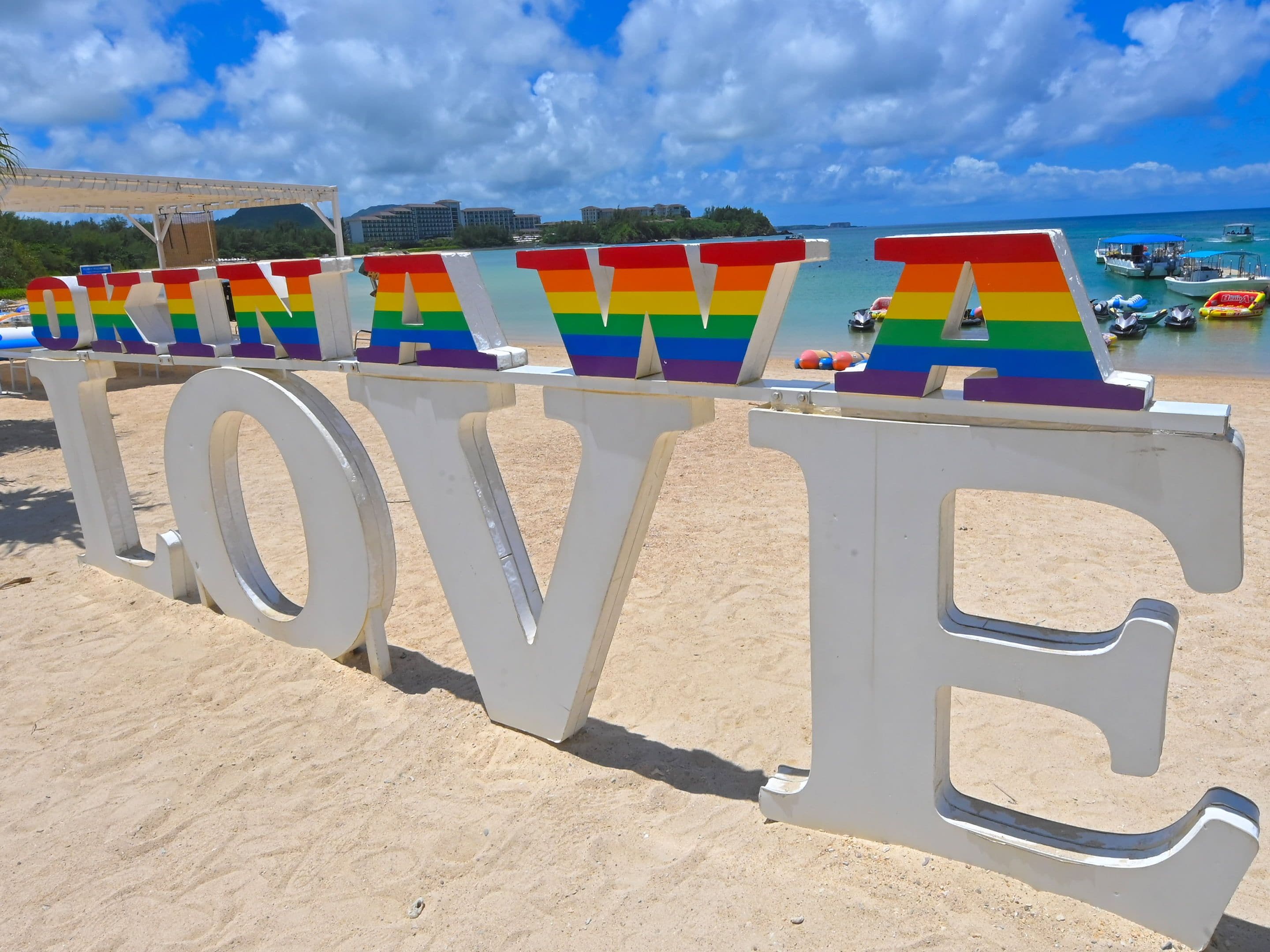Una playa con letras blancas grandes que deletrean 'LOVE', con vistas a barcos y edificios al fondo.