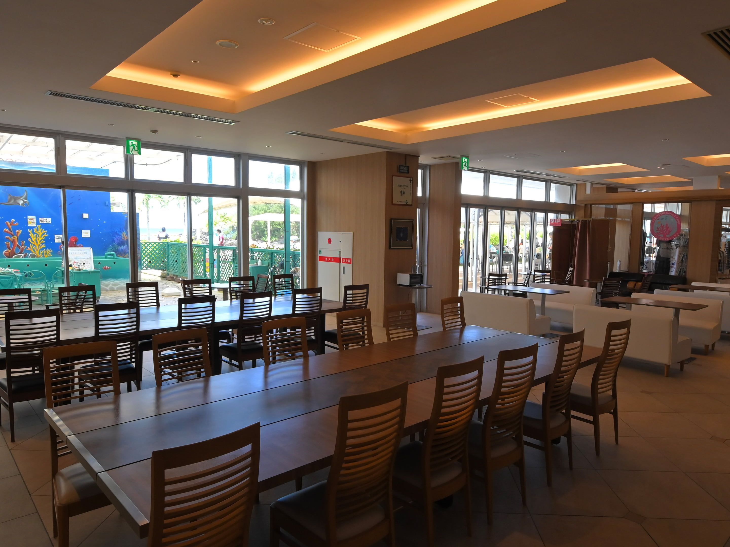 Un comedor amplio con varias mesas y sillas, ventanas grandes que ofrecen vistas del exterior, y una recepción.