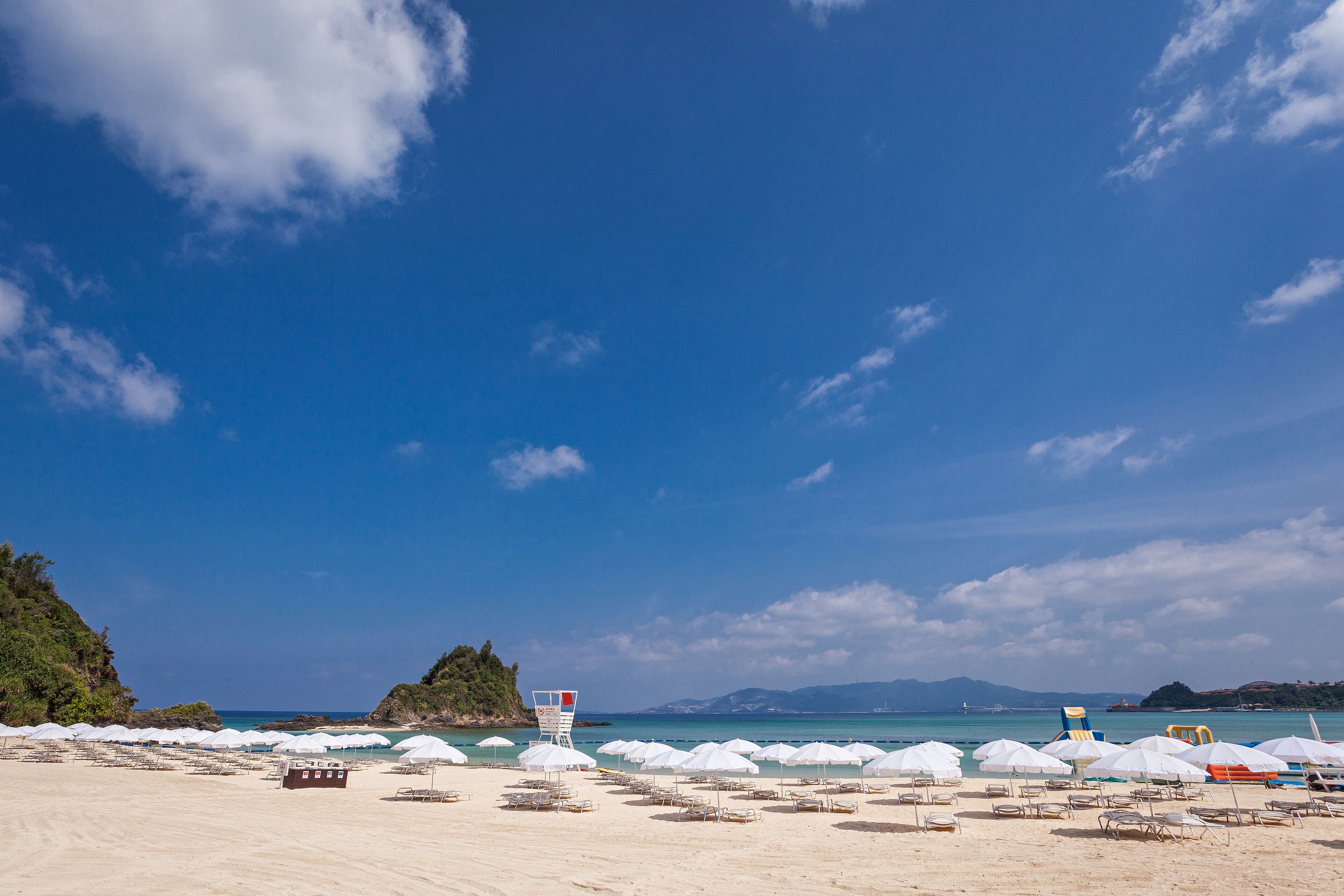 Ein Strand mit weißen Sonnenschirmen und Liegestühlen, einem Rettungsturm und blauem Himmel.
