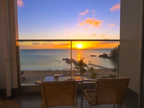 Un balcon agrémenté de fauteuils en osier, d’où l’on peut admirer un magnifique coucher de soleil sur la mer, un quai et une côte rocheuse.