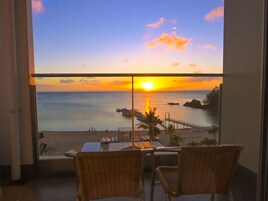 A balcony with wicker chairs overlooking a sunset view of the sea, a pier, and a rocky coastline.