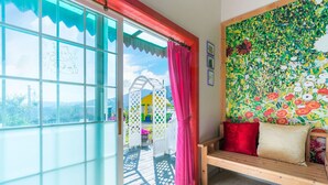 Een kamer met een houten bankje, een kleurrijke bloemenwand en uitzicht door een glazen deur.