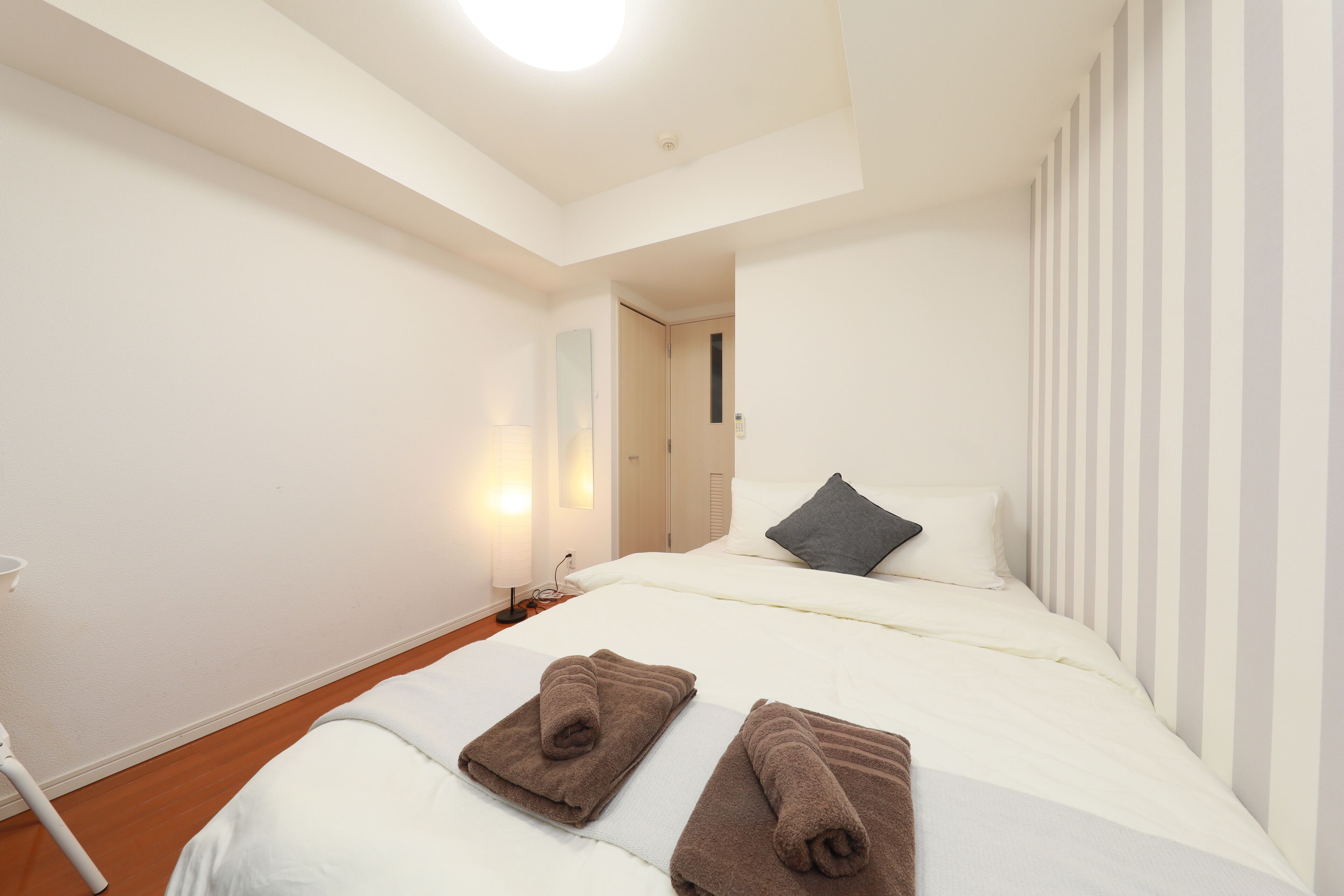 A hotel room with a bed, two towels, a nightstand, a lamp, and a door leading to another room.