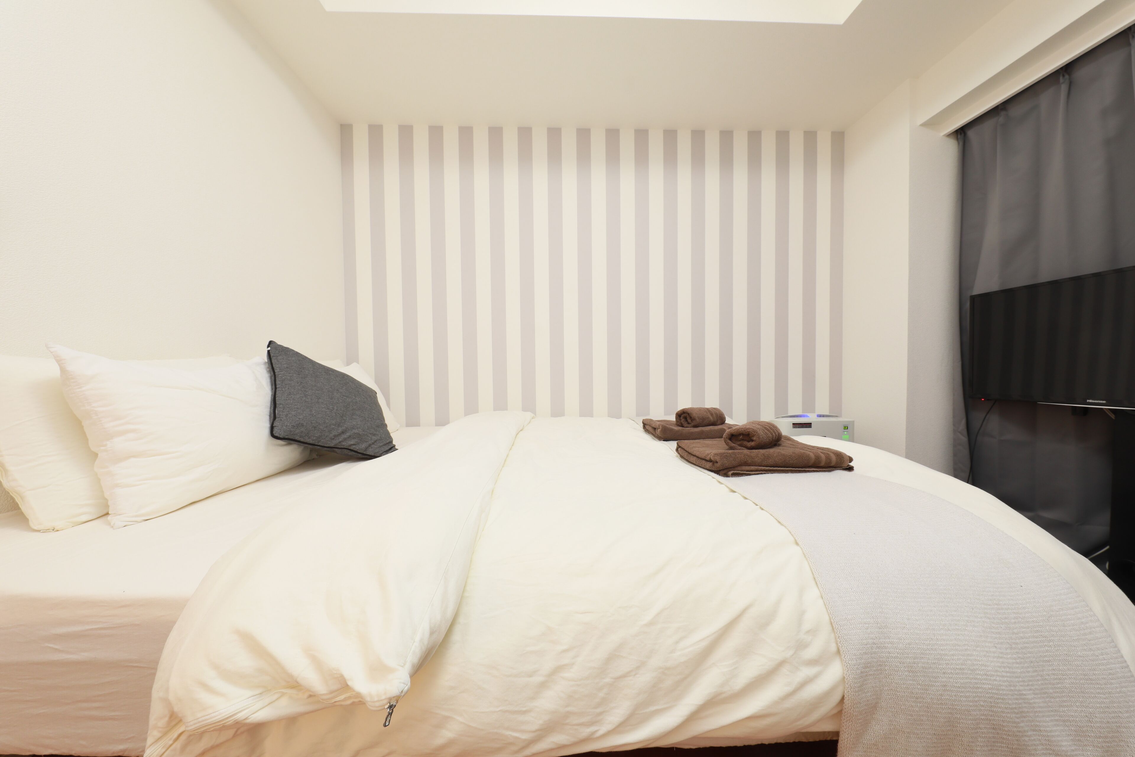 Een net opgemaakt bed met kussens, een dekbed en een paar slippers. Er is een raam met zonwering en een televisie aan de muur.